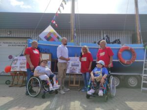 Dwie osoby na wózkach inwalidzkich Za nimi stojące osoby w czerwonych koszulkach na tle niebieskiego kadłuba łodzi