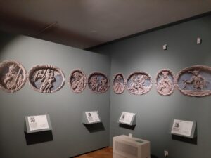 Kolorowe fotografie kamiennych medalionów ściennych z opisami umieszczone na szarej ścianie