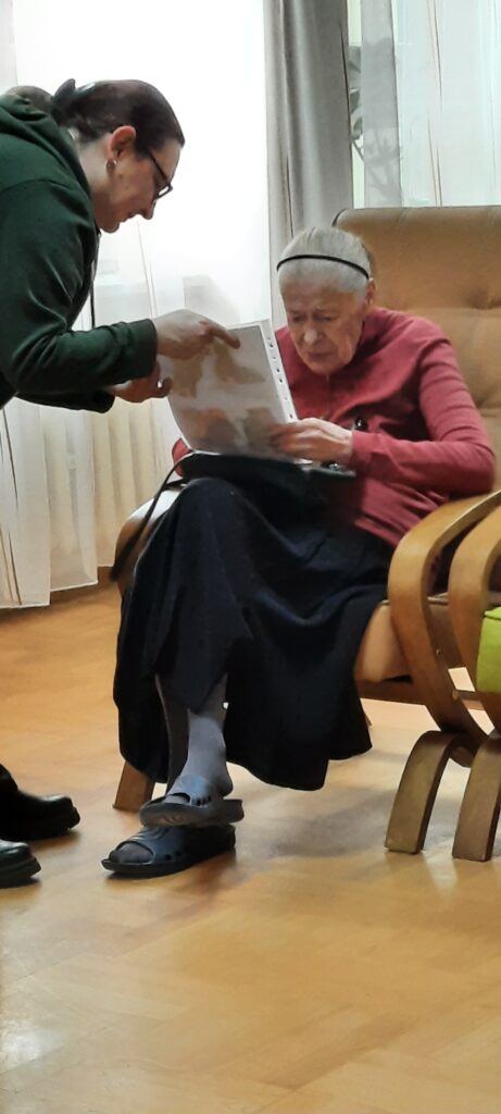 Kobieta w okularach pochyla się i podaje kartkę kobiecie siedzącej w brązowym fotelu