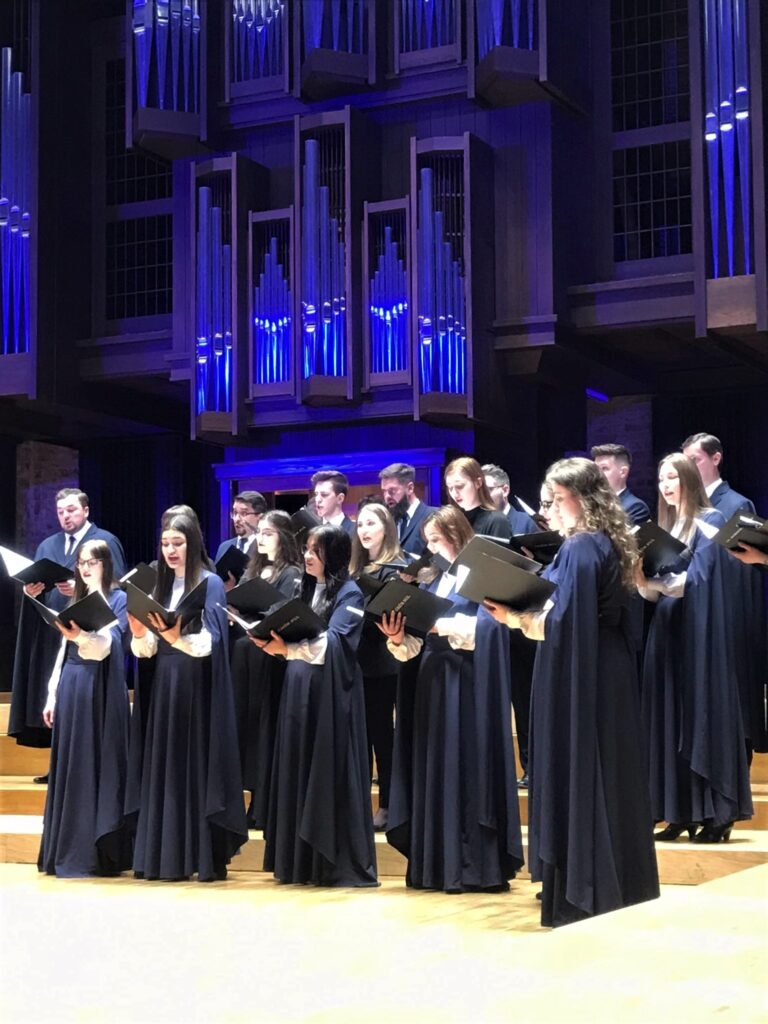 Liczni członkowie chóru ubrani w czarne peleryny i stojący na tle oświetlonych na niebiesko organów