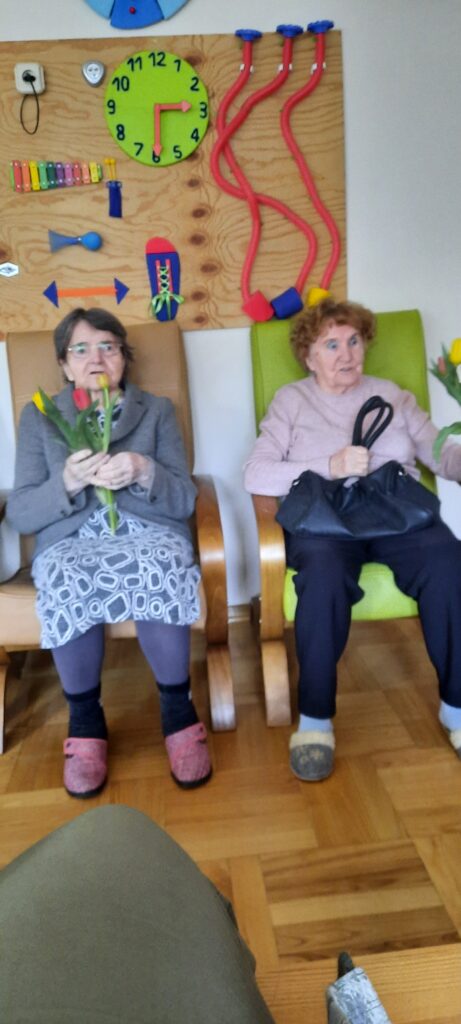 Kobiety w fotelach - jedna trzyma czarną torebkę a druga wiązankę żółtych tulipanów