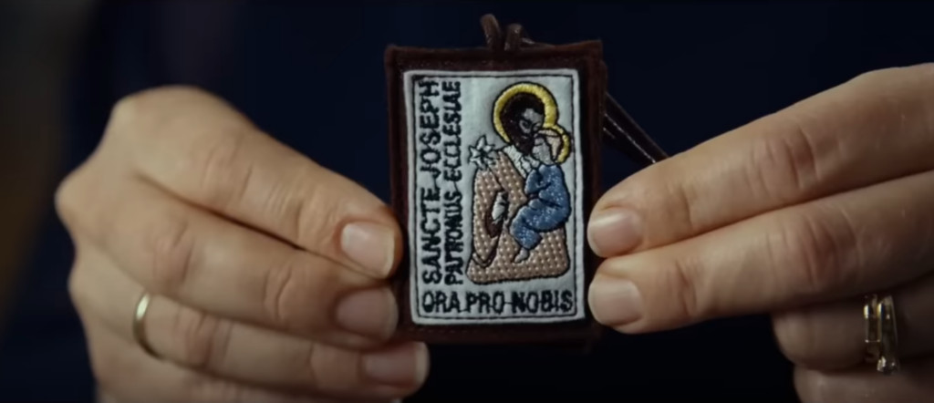 Kolorowo wyszywany szkaplerz ze świętym Józefem trzymany w palcach obydwu dłoni