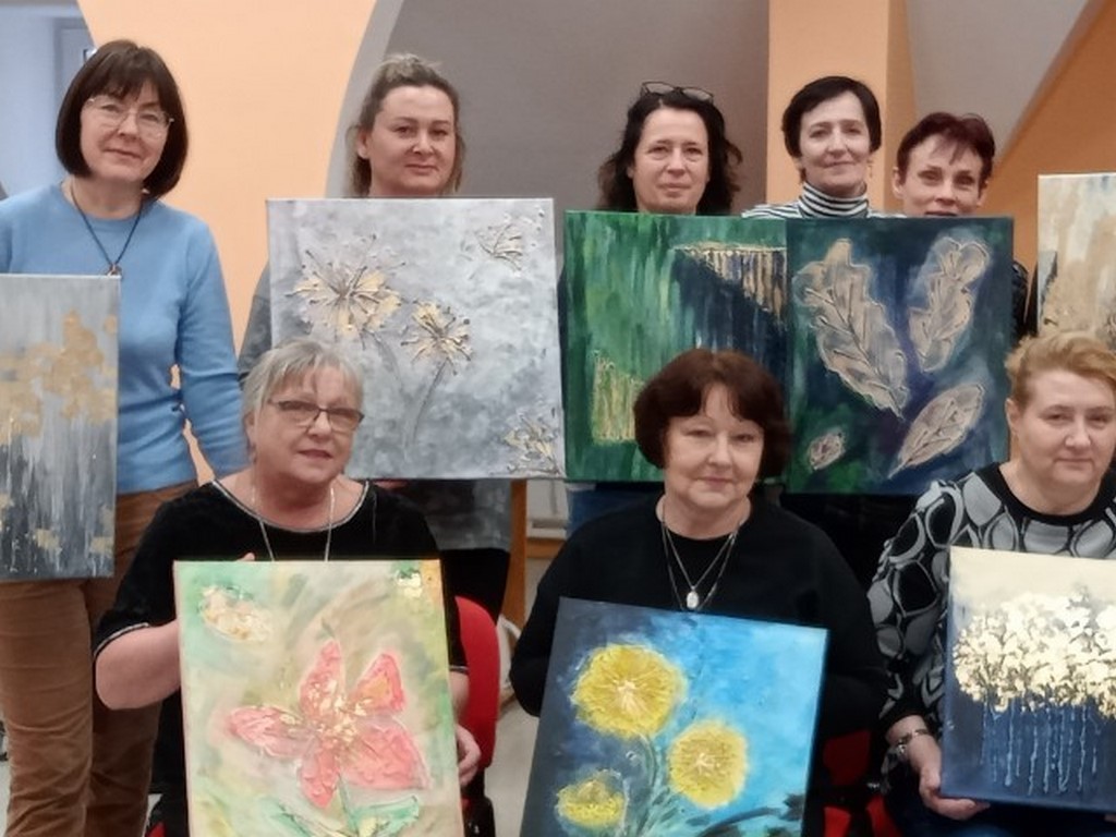 Osiem stojących kobiet trzymających przed sobą kolorowe prace plastyczne