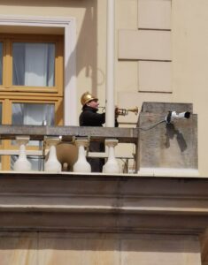 Człowiek w strażackim mundurze stoi na kamiennym balkonie i gra na trąbce