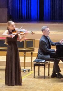 Kobieta w długiej brązowej sukni grająca na skrzypcach i mężczyzna w czarnym garniturze siedzący przy fortepianie