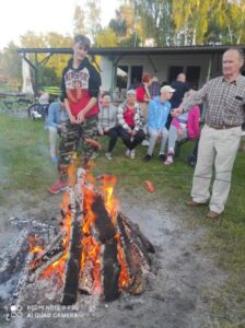 Grupa osób trzymająca długie kijki z kiełbaskami i stojąca przy ognisku