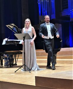 Kobieta w jasnej sukni i mężczyzna we fraku stojący na tle fortepianu i widocznych w tle organów oświetlonych na niebiesko
