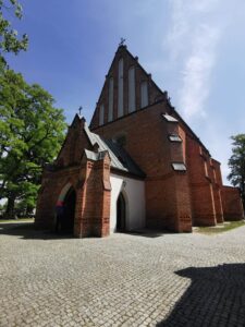 Niewielki gotycki kościół z czerwonej cegły widoczny od strony wejścia i otoczony brukowanym placem