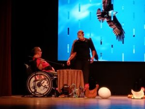Stojąca osoba w stroju orła i osoba na wózku inwalidzkim przy stoliku nakrytym obrusem