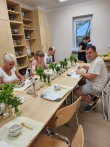 Cztery osoby siedzące przy stole zastawionym zielonymi wiązankami ziół w przeźroczystych wazonach
