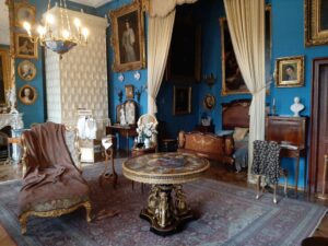 Pałacowe wnętrze z niebieskimi ścianami ozdonym stołem piecem kaflowym kamiennymi popiersiami i obrazami
