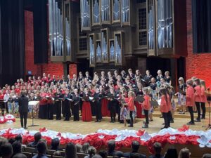 Duża grupa osób ze śpiewnikami stojąca na scenie na tle organów