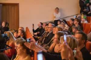Publiczność widziana z lewej strony siedząca w pomarańczowych fotelach na sali widowiskowej
