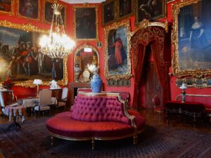 Sala pałacowa z meblami wypoczynkowymi na środku i licznymi obrazami na czerwonych ścianach