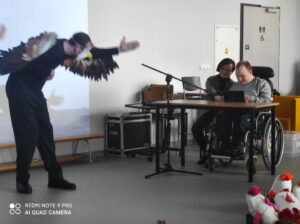 Stojąca osoba ze skrzydłami orła i wyciągnietą do przodu ręką oraz siedzące obok dwie osoby przy stoliku
