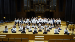 Grupa kilkudziesięciu dziewczyn i chłopców ubranych w czarno-białe stroje i  znajdujących się na dużej drewnianej scenie