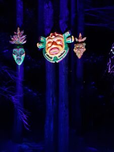 Iluminacja świetlna w formie trzech masek totemowych zawieszonych oddzielnie na pniach drzew