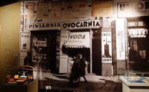 Kilkumetrowa fotografia ulicy z wejściami do sklepów i szyldami z okresu początku XX wieku naklejona na ścianie oraz dwie gabloty z artykułami przemysłowymi