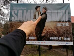 Lewa dłoń z wystawionym palcem wskazującym na tle billboardu reklamującego film Sami Swoi początek
