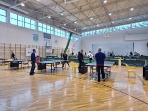 Hala sportowa z rozstawionymi zielonymi stołami i zawodnicy grający w tenisa stołowego
