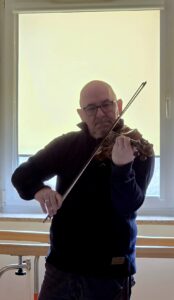 Łysy mężczyzna w okularach grający na skrzypcach na tle okna zasłoniętego jasna roletą