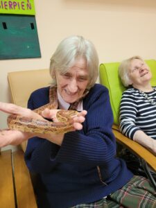 Dwie starsze kobiety siedzące w fotelach i jedna z nich trzyma żywego węża w dłoniach
