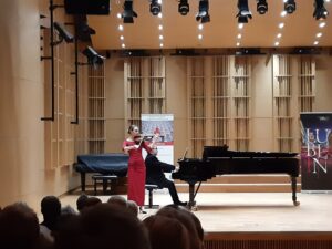 Postaci na drewnianej scenie - kobieta w czerwonej sukience grająca na skrzypcach oraz mężczyzna w garniturze akompaniujący na fortepianie