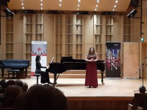Postaci na drewnianej scenie - śpiewająca kobieta w czerwonej sukience oraz kobieta w czarnej sukience akompaniująca na fortepianie