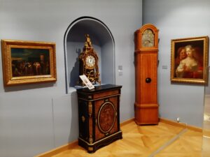 Mosiężny zegar eksponowany w ściennej wnęce, duży zegar stojący, kredens oraz dwa obrazy olejne w drewnianych ramach
