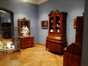 Sala muzealna z zabytkowymi kredensami, białą porcelaną, niewielkimi formami rzeźbiarskimi oraz obrazami olejnymi
