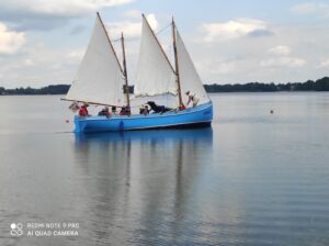 Trójmasztowa łódź z niebieskim kadłubem wypełniona ludźmi i płynąca po jeziorze