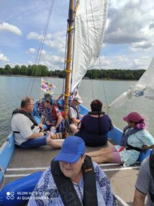 Grupa dorosłych osób w nakryciach głowy siedząca na niebieskim pokładzie łodzi z żaglami