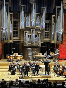 Na pierwszym planie widoczne głowy widowni i fotele natomiast dalej widać orkiestrę z dyrygentem a tło wypełniają duże organy zajmujące całą ścianę filharmonii