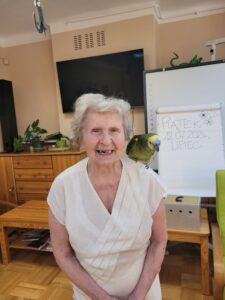 Starsza kobieta z zielonożółtą papugą na lewym ramieniu stojąca na tle telewizora i ubrana w białą bluzkę