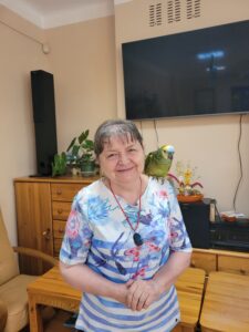 Starsza kobieta z zielonożółtą papugą na lewym ramieniu stojąca w pomieszczeniu i ubrana w jasną bluzkę