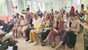 Duża grupa dorosłych osób siedzących na krzesłach w kilku rzędach na tle okien