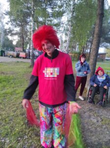 Uśmiechnięty mężczyzna ubrany w kolorowe ubrania i czerwoną perukę z prostymi włosami stojący na tle zielonych drzew