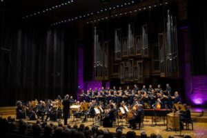 Drewniana scena w filharmonii z orkiestrą i dyrygentem oraz widocznymi w tle organami na całą ścianę oświetlonymi fioletowymi odcieniami