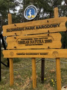 Drewniana tablica z desek z czarnymi literami z informacją o Słowińskim Parku Narodowym oraz kolorowe logo nad nimi