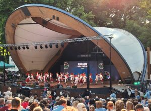 Duża grupa dziewczynek w kolorowych strojach ludowych tańczących w parach na drewnianej scenie w muszli koncertowej