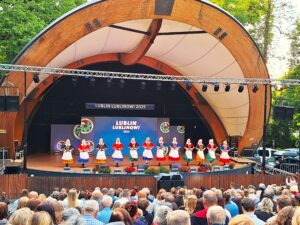 Dwanaście kobiet w kolorowych strojach ludowych stojących w rzędzie na drewnianej scenie w muszli koncertowej