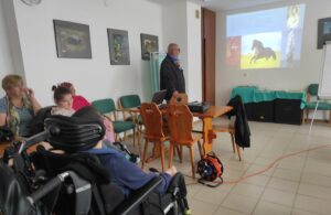 Grupa osób siedząca przy drewnianym stole i patrząca w kierunku mężczyzny stojącego przy projekcji wyświetlonej na ścianie
