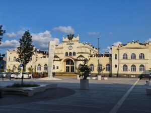 Jasnożółty budynek Dworca Kolejowego Lublin Główny oraz betonowy plac przed nim z pojedynczymi drzewkami w donicach oraz zegarem umieszczonym na pionowej formie z betonu