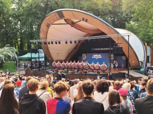 Kobiety w kolorowych strojach ludowych i pasiastych spódnicach stojące w rzędzie na drewnianej scenie w muszli koncertowej