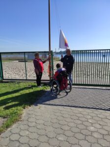 Osoba na wózku inwalidzkim i dwie inne stojące przy maszcie podczas pociągania kolorowej bandery