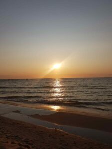 Płaszczyzna morza z fragmentem piaskowej plaży oświetlona światłem zachodzącego słońca odbijającego się też w wodzie