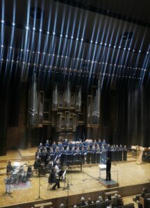 Wnętrze oświetlonej sceny w filharmonii z chórem i grającymi muzykami oraz widocznymi organami w tle