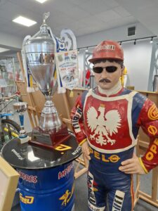Manekin mężczyzny z namalowanym na nim kombinezonie żużlowca oraz duży srebrny puchar ustawiony na okrągłym blacie umieszczonym na niebieskiej metalowej beczce