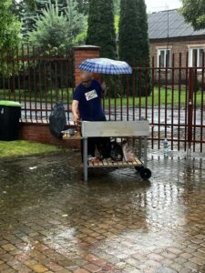 Mężczyzna w niebieskiej koszulce trzymający rozłożoną parasolkę i stojąc przy grillu ustawionym na mokrej kostce brukowej