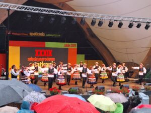 Na pierwszym planie duża grupa osób z otwartymi parasolkami a w tle tańczące na scenie pary w strojach ludowych 